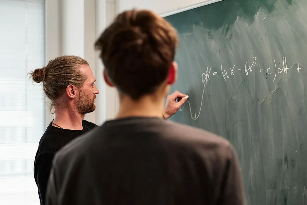 Zwei Männer vor einer grünen Tafel, einer von ihnen schreibt mit Kreide eine mathematische Formel