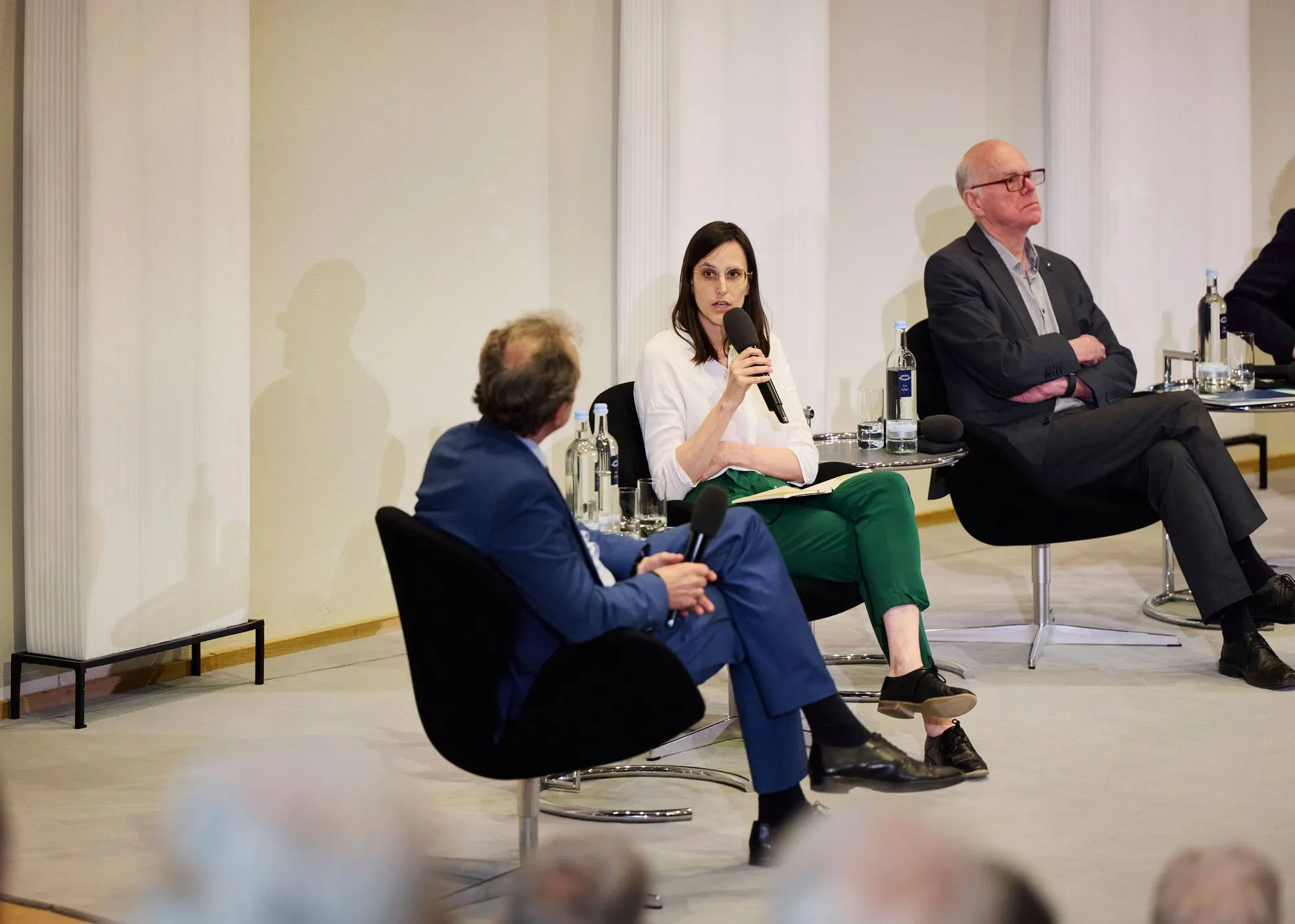 Frau sitzt auf einem Podium zwischen zwei Männern und spricht in ein Mikro