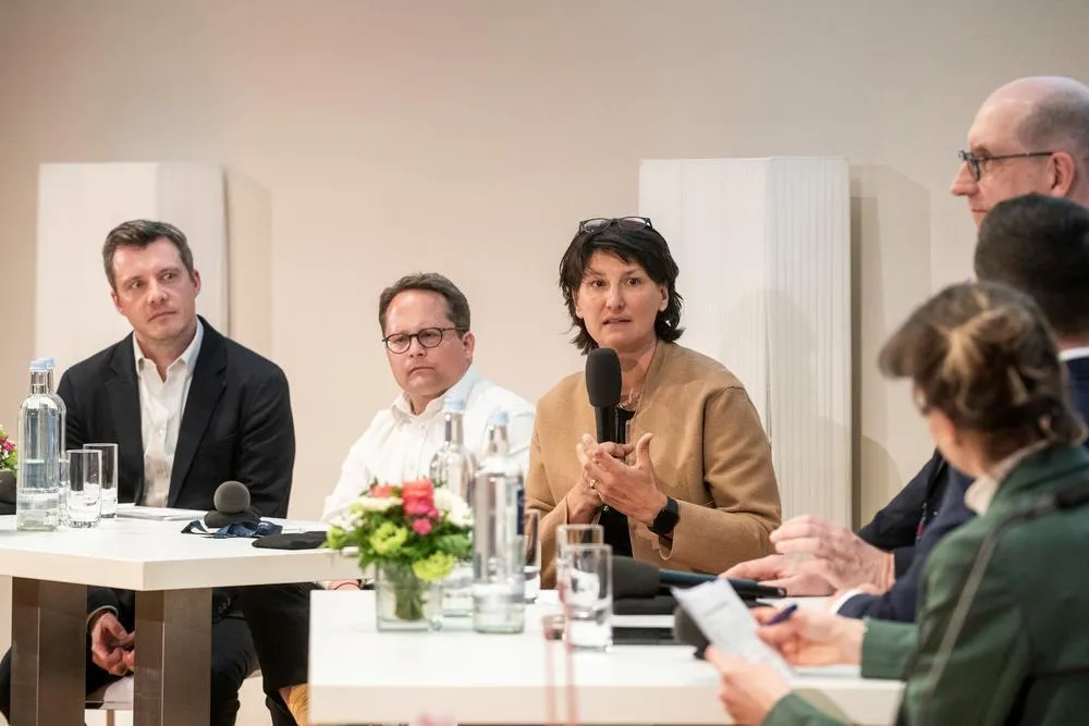 Eine Frau spricht in ein Mikro, fünf andere Menschen hören ihr zu