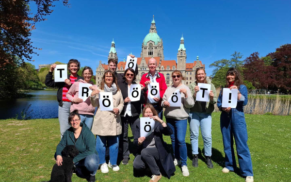 Eine Gruppe von Personen vor dem Neuen Rathaus Hannover, alle halten Zettel mit Buchstaben hoch, die zusammen das Wort Trööööööt bilden
