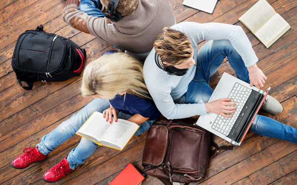 Das Foto zeigt zwei Personen von oben, wovon eine Person ein Buch liest und die andere am Laptop arbeitet.