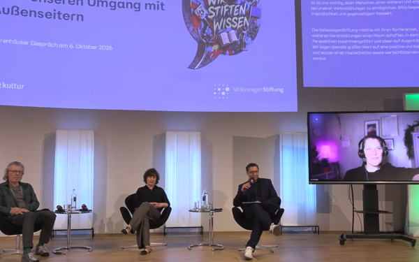 Drei Personen sitzen auf einem Podium und eine weitere ist vie Bildschirm zugeschaltet.