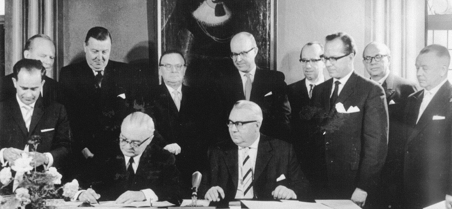 Mehrere Männer in dunklen Anzügen stehen um einen Tisch, an dem zwei Männer Unterlagen unterzeichnen