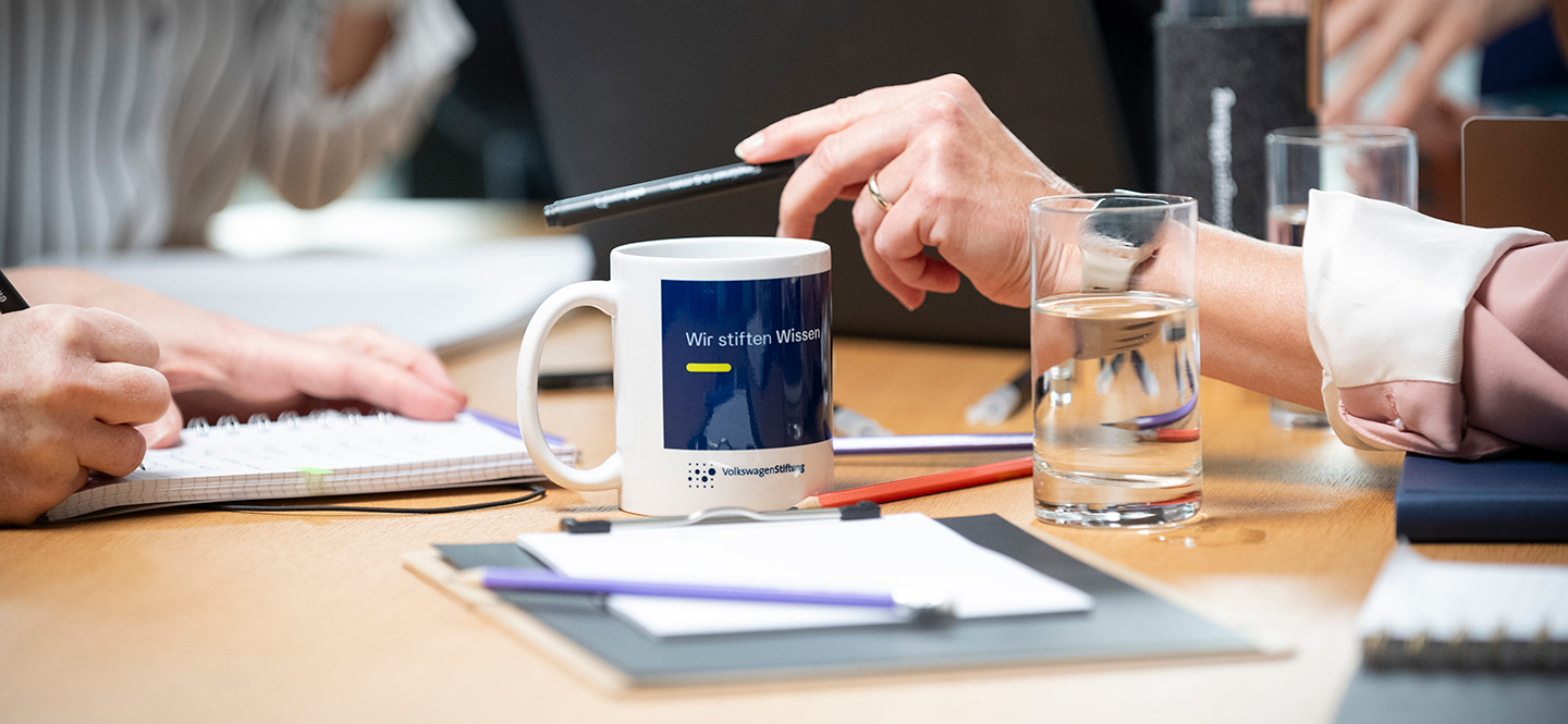Arbeitssituation mit mehreren Personen an einem Tisch, zu sehen sind Schreibutensilien, eine Tasse, Hände