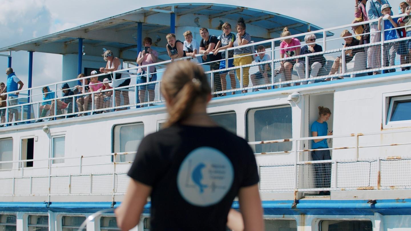 Frau steht vor einem Schiff, auf dem Zuschauer stehen.