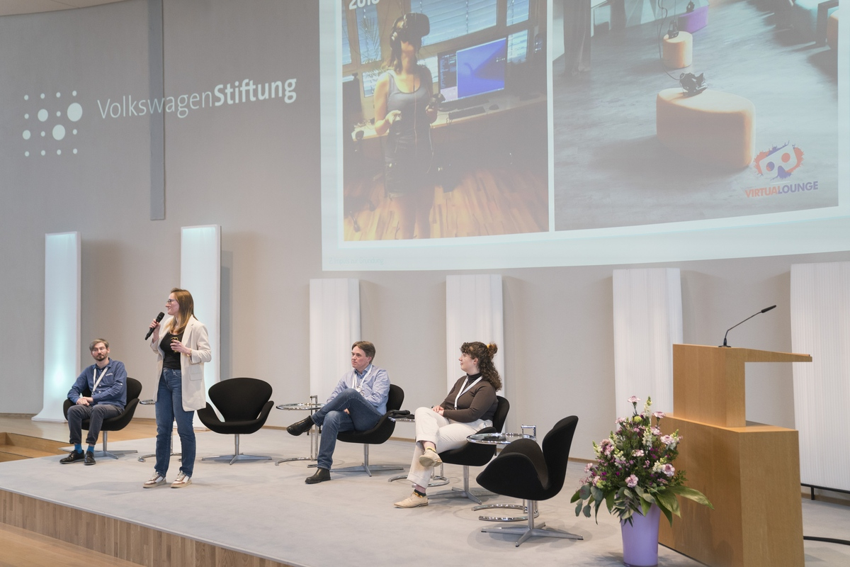 Eine Frau präsentiert etwas auf dem Podium, zwei Männer und eine weitere Frau sitzen daneben 