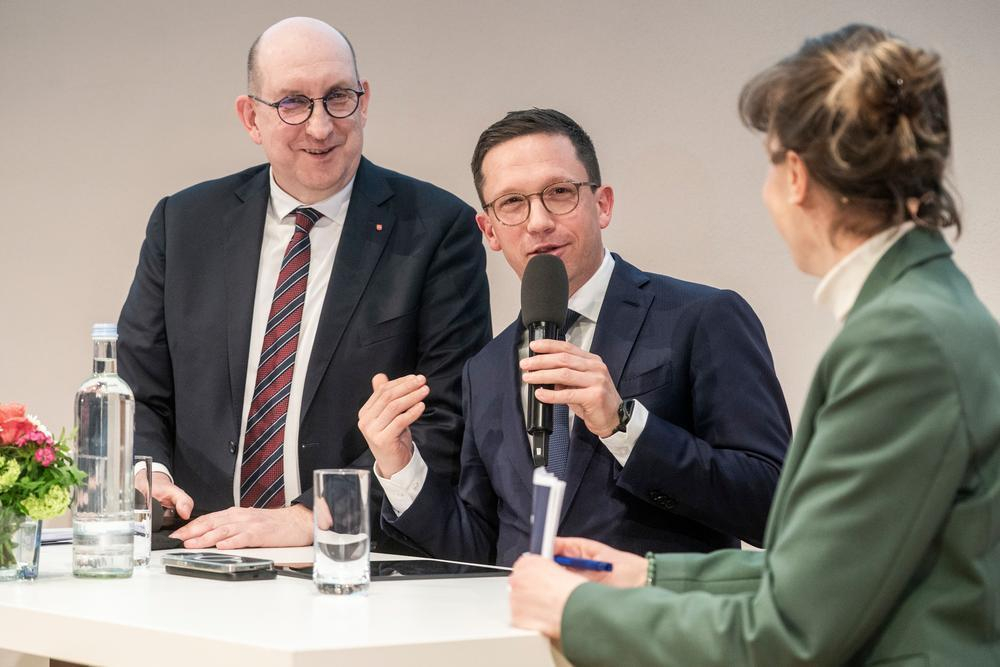 Zwei Männer, einer davon spricht in ein Mikrofon, stehen neben einer Frau