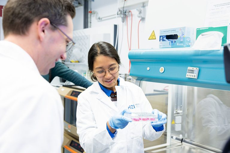Eine Frau im Kittel vor einem Abzug, in der Hand eine Petrischale