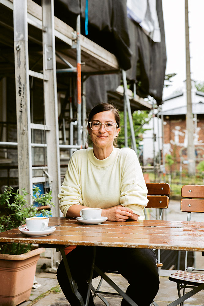 Frau sitzt mit einem Kaffee draußen am Tisch und lächelt in die Kamera 