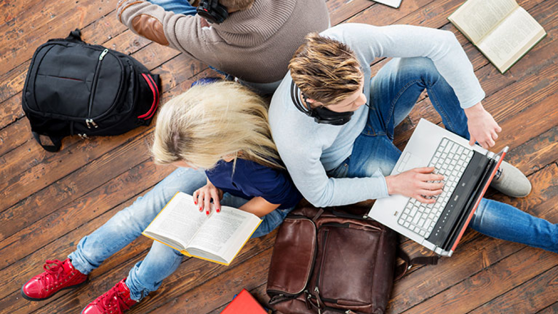 Das Bild zeigt von oben drei sitzende Personen. Eine liest ein Buch, die andere hat einen Laptop auf dem Schoß.