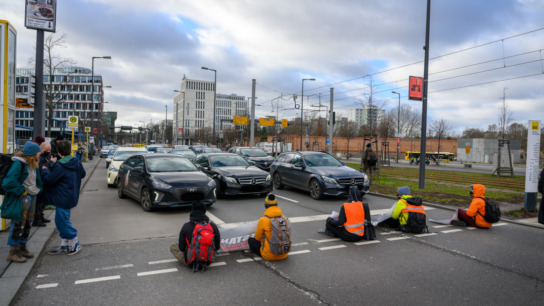 Das Bild zeigt Menschen, die auf einer Straße sitzen und den Verkehr blockieren.