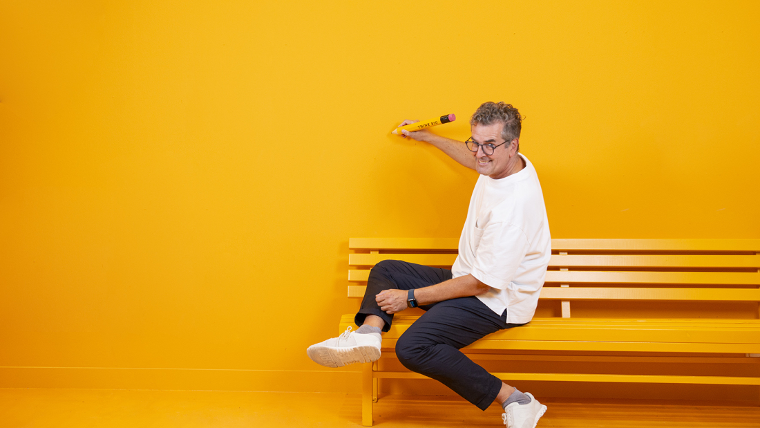 Das Bild zeigt Gunnar Spellmeyer mit einem kreativen Stift auf einer Bank vor einer gelben Wand sitzend und schreiben